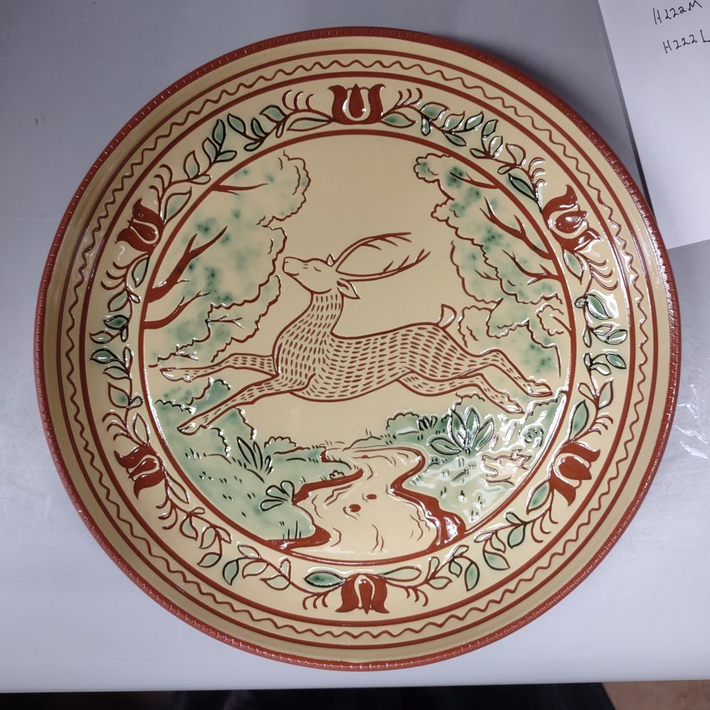A photograph of a redware Sgraffito plate with a drawing of a leaping deer in the center. The deer is jumping across a small river, green foliage is visible on either riverbank. In the bottom right is a barking dog head, indicating that the deer is being chased. The border has a repeating floral tulip design. Soft greens accent the plant decorations on the plate. 