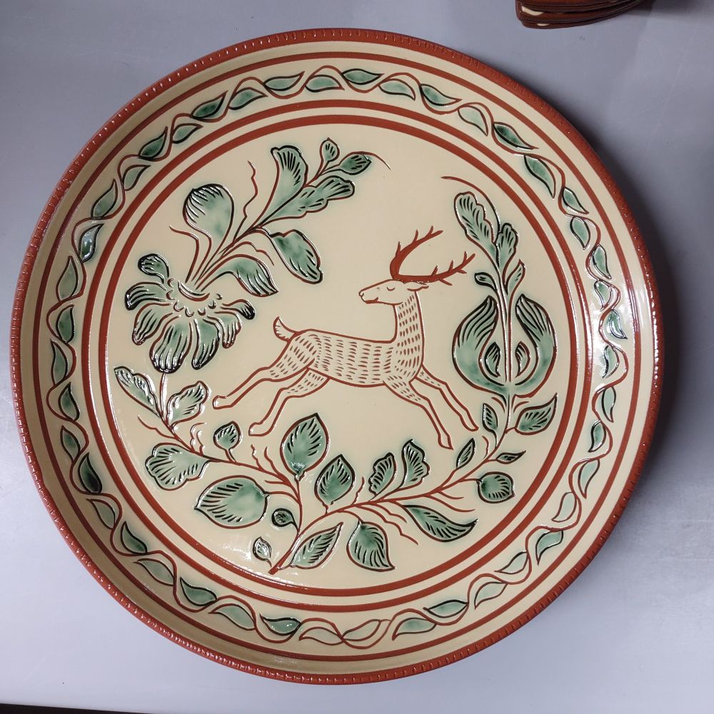 Another redware plate with a sgraffito design of a deer. The deer is leaping to to right, and surrounded by two large branching plants. The border of the plate has an alternating leaf pattern. The plants on the plate are accented with a soft green. 