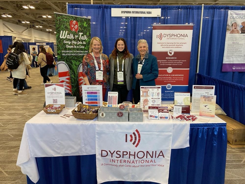 Three women standing in front of a table containing information and resources on dysphonia