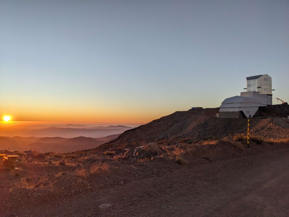 A sunset view over clouded mountains, with the Vera C. Rubin Observatory building on the right
