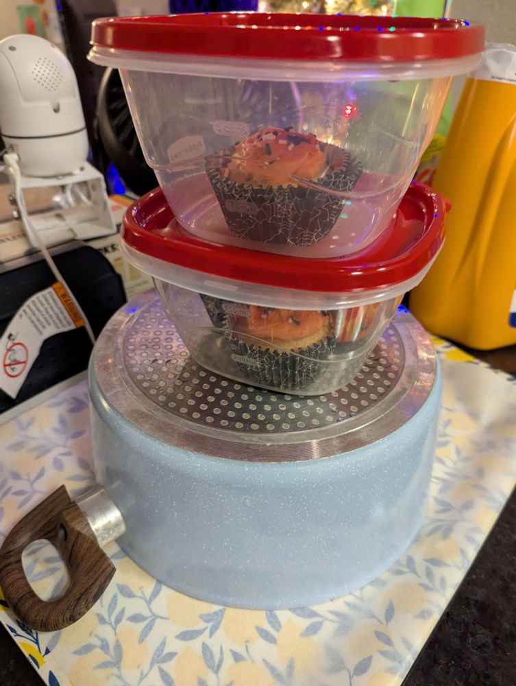 A stack of kitchenware. A large pot on the bottom that is flipped upside down on a serving tray. On top of it are two smaller plastic Gladware containers containing cupcakes . 