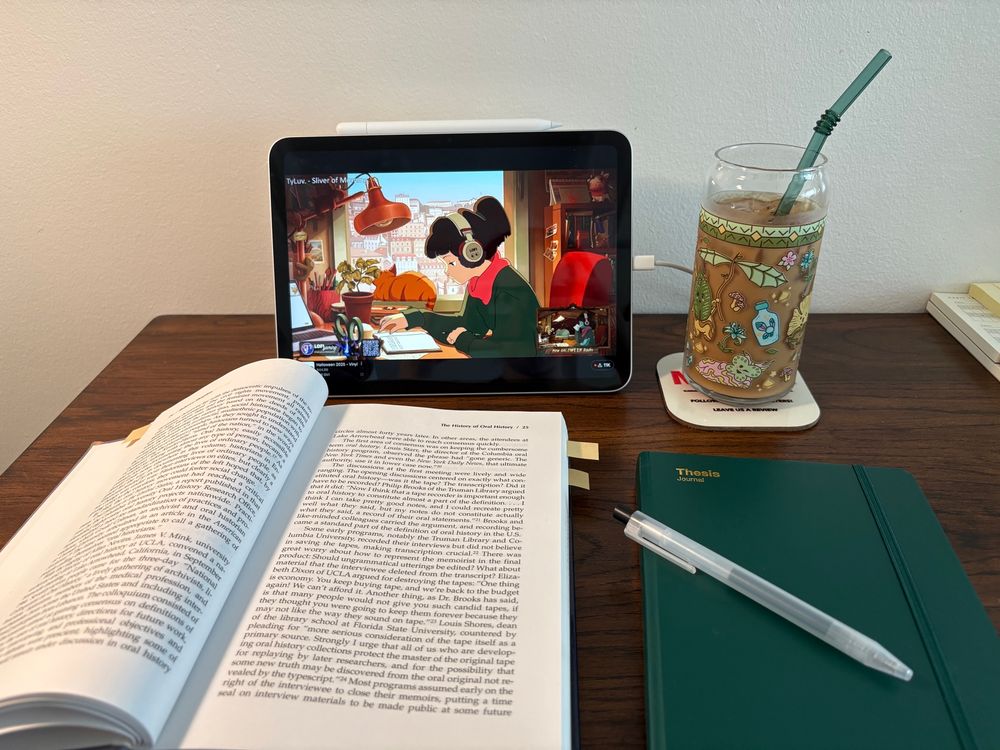 My desk setup with a book, my thesis journal, an iced coffee, and lofi girl playing on my iPad 