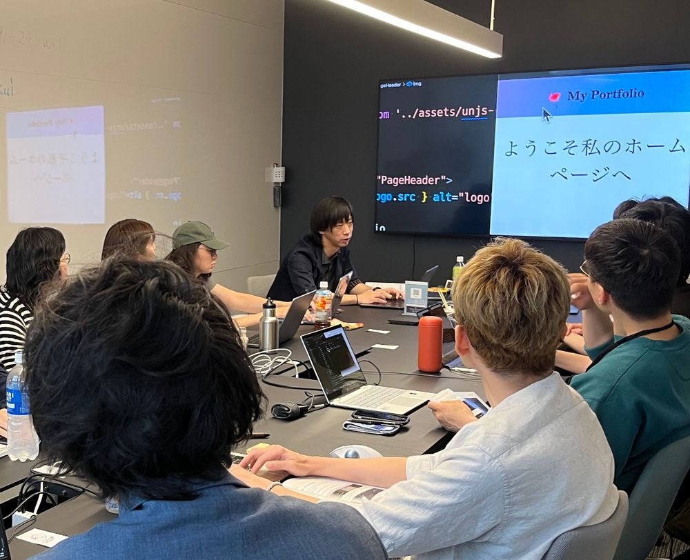 CodeGridワークショップの様子。会議室のような場所で、1つの机に向かい合って講師1名と参加者が7名並んで座っている。壁にはモニタがあり、コードやWebサイトが表示あれている。