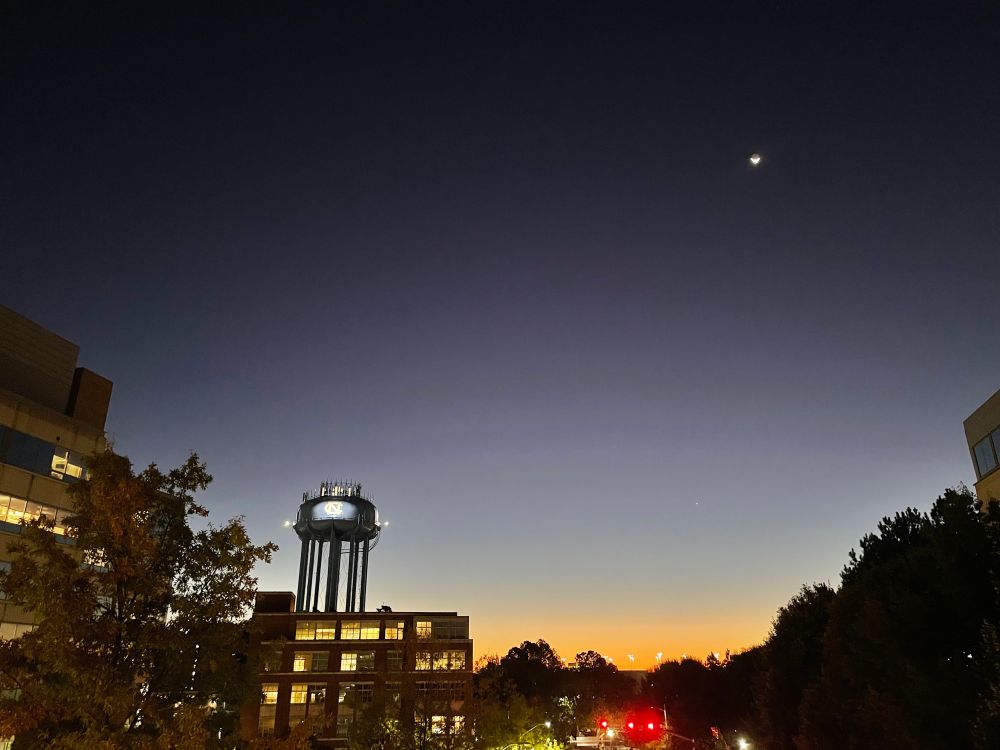 Photo of predawn just lighting the horizon. You can see a tiny sliver of a moon in the dark sky. In the mid-ground is the UNC (University of North Carolina) water tower and some buildings. 
