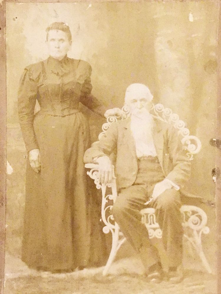 Very old photo of and old man with a long, white beard sitting in a wicker chair. Standing beside him is an older woman in a severe black dress. 