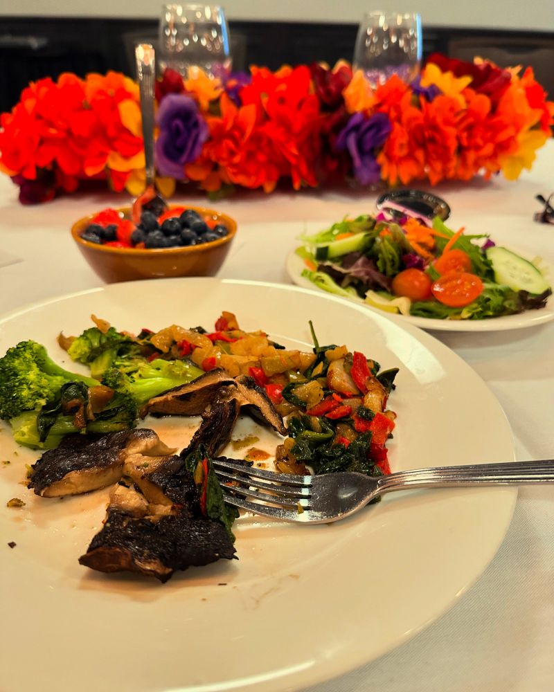 Three plant-based dishes at a conference table: a bowl of strawberries and blueberries, a salad plate, and portobello mushroom steak as the main dish.