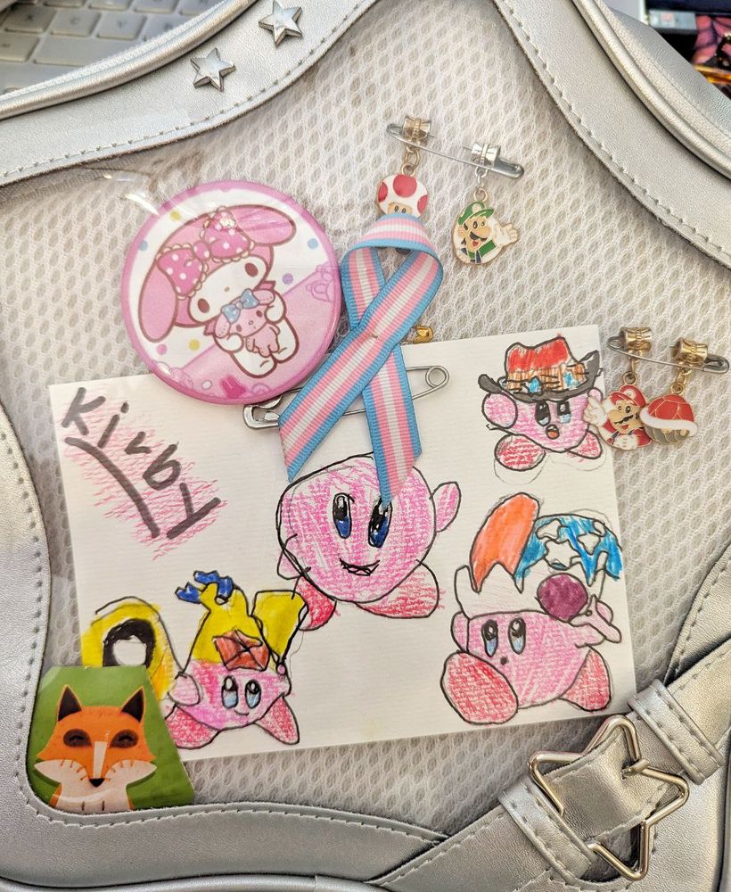 
Blick auf einen sternenförmigen Rucksack aus silbernem Kunstleder. Die Front besteht aus dicker Klarsichtfolie, darunter ist ein mit Kinderhand gemaltes Bild mit mehreren Kirbys zu sehen, ein Schleife in den Farben der Transflagge, ein My Melody Button und 2 Sicherheitsnadeln, von denen jeweils Super Mario, Luigi, Toad und ein roter Panzer baumeln.