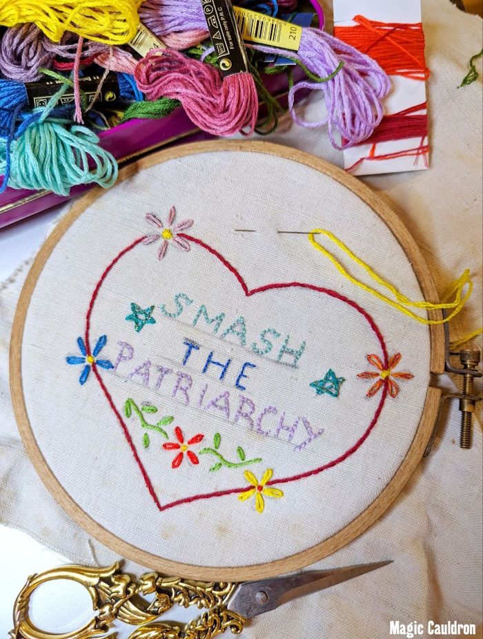 A  wooden embroidery hoop with old,  thick, handwoven linnen. Above is a tin full of colourful embroidery floss. Beneath ist a golden, flowery looking embroidery scissor. The embroidery: a big red heart with 4 simple flowers crossing the red embroidery. Inside in turquoise, blue violet ist the text "SMASH THE PATRIARCHY" embroidered, flanked by stars. Underneath is another red flower with green leaves. A needle with yellow thread is woven though the fabric. The work seems to be finished recently, you can still see some of the pencil lines of the sketch for the embroidery.