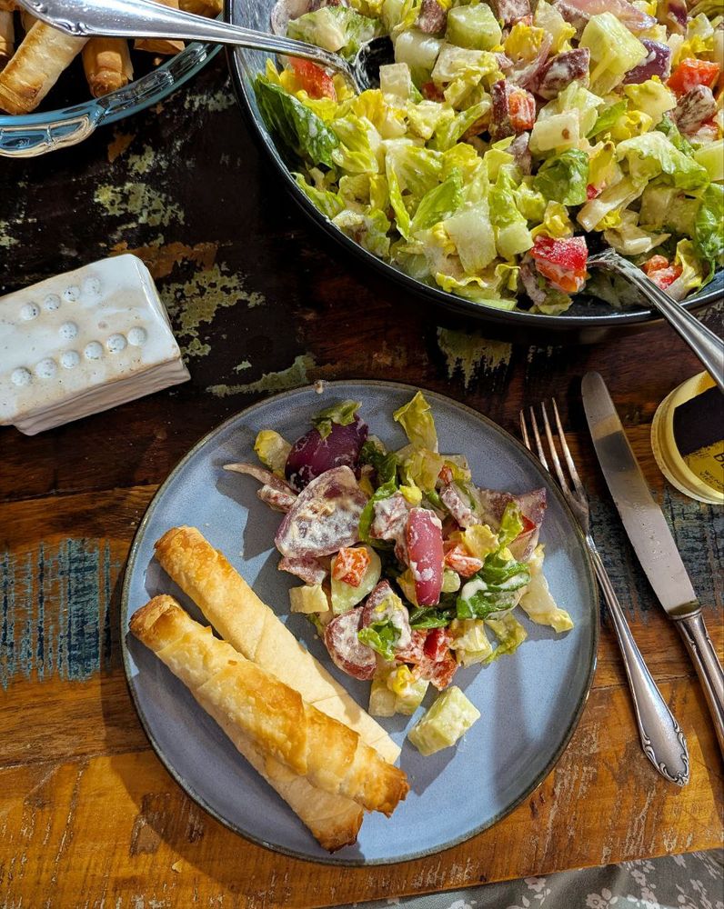 Dunkler Tisch. Darauf ein Abendbrotteller auf dem 3 braun gebackene Zigarrenbörek liegen, daneben ein Haufen Salat mit weißem Dressing. Rundherum auf dem Tisch 1 weiße Porzellandose für Salz, 1 Glasschale mit Börek, Besteck, 1 große Schale mit Salat, 1 Limoflasche.