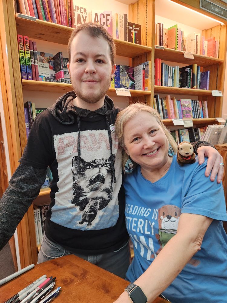 A young caucasion male in a shirt that is a pun of JAWS but its a cat that says PAWS, next to Katherine Applegate who is wearing an otter shirt to promote her picturebook "Odder". You can't see it but shes got on Otter slippers. 