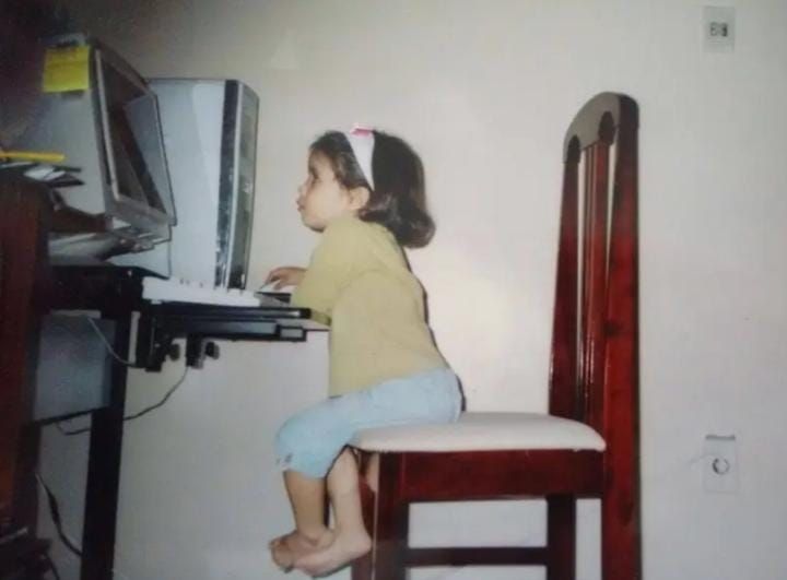 uma criança de cabelo curto chanel e tiara está sentada numa cadeira de madeira 2x maior que ela,  na ponta, para usar um computador desktop doa anos 2000 com monitorzão de tubo. ela parece muito concentrada 