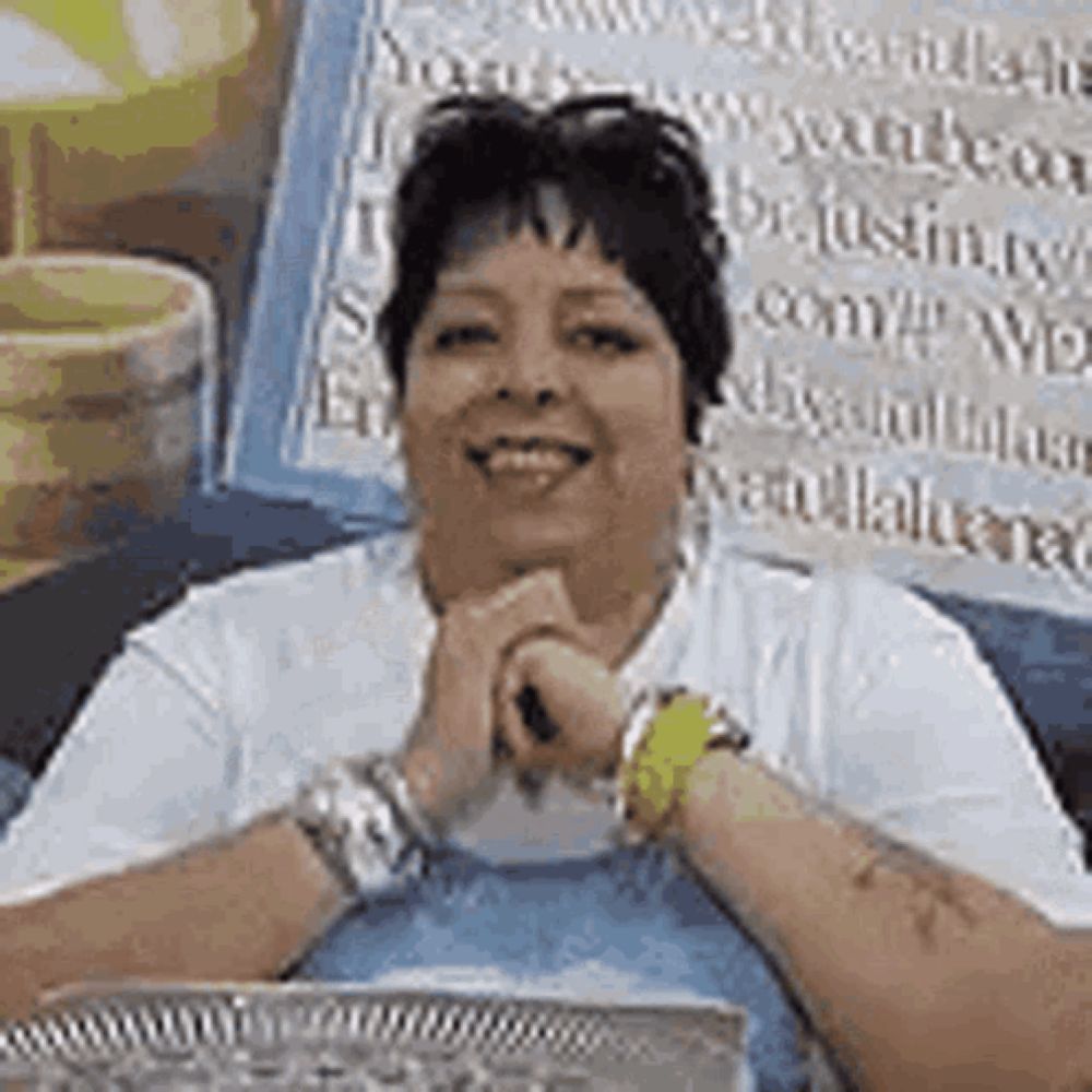 a woman is sitting at a table with her hands folded in front of her face and smiling .