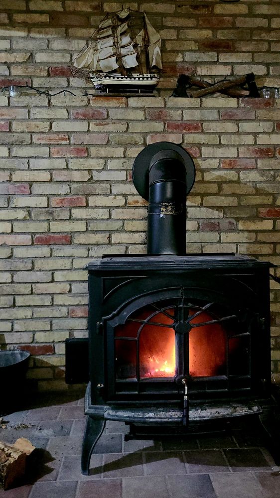 A fire is ablaze inside a black cast iron wood stove. The stove rests on an earth-toned tile floor in front of a worn, light-toned brick chimney. A model ship, two antique axes, and string lights decorate the mantle. A lone pine log and an ash can peek into the frame, sitting on the tile to the left side of the stove.