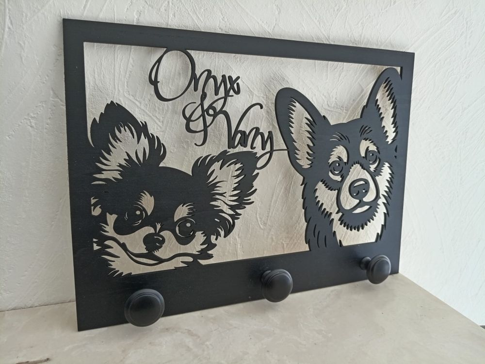 Porte-laisses mural en bois noir découpé au laser, représentant les portraits de deux chiens de type chihuahua et corgi avec les prénoms "Onyx & Vany" inscrits en haut. Trois patères noires sont fixées en bas pour accrocher les accessoires.
