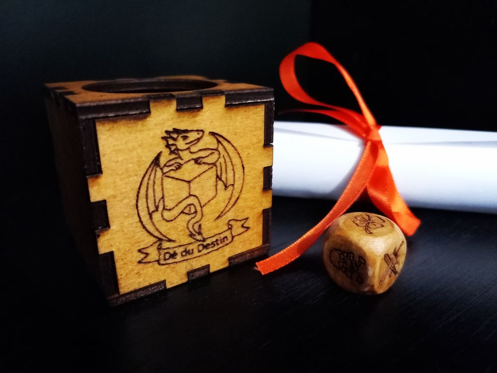 Une boîte en bois orné d'un dragon qui tient un d6, avec une bannière "dé du destin", avec un dé 6 en bois gravé et un parchemin roulé au ruban orange. 