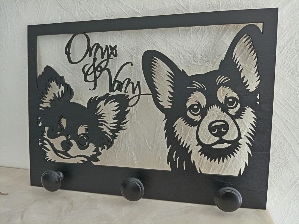 Porte-laisses mural en bois noir découpé au laser, représentant les portraits de deux chiens de type chihuahua et corgi avec les prénoms "Onyx & Vany" inscrits en haut. Trois patères noires sont fixées en bas pour accrocher les accessoires.