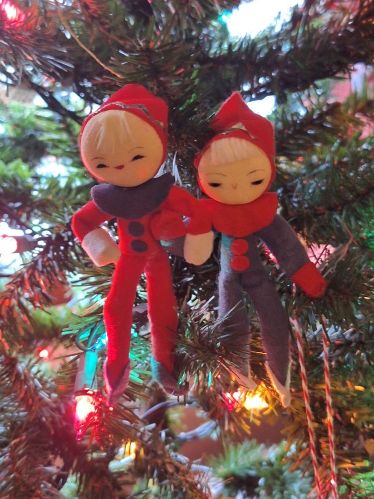 2 vintage elf dolls. One in red felt with green trim, the other in green with red.
They are holding hands, hanging from a Christmas tree with colored lights in the background.