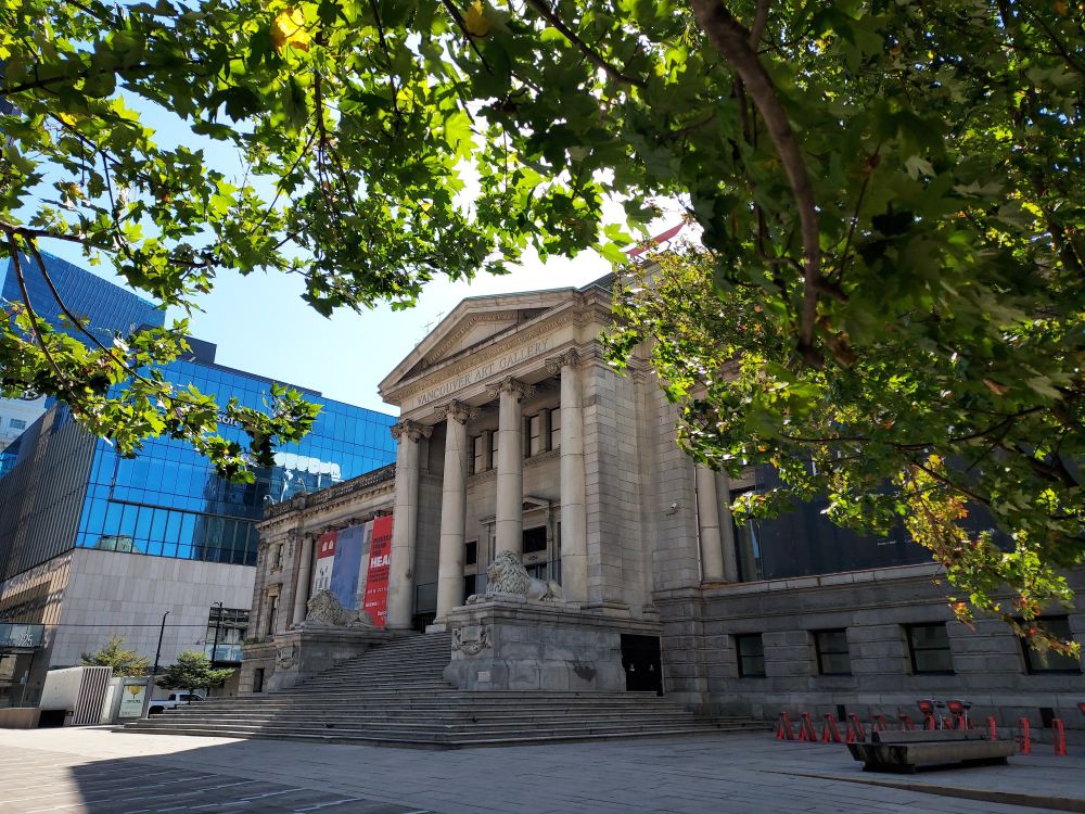 A view, looking from under a tree, of the neoclassical facade of the Vancouver Art Gallery. It has four tall columns and a triangular pediment. There are leaves and branches framing the view at the top.