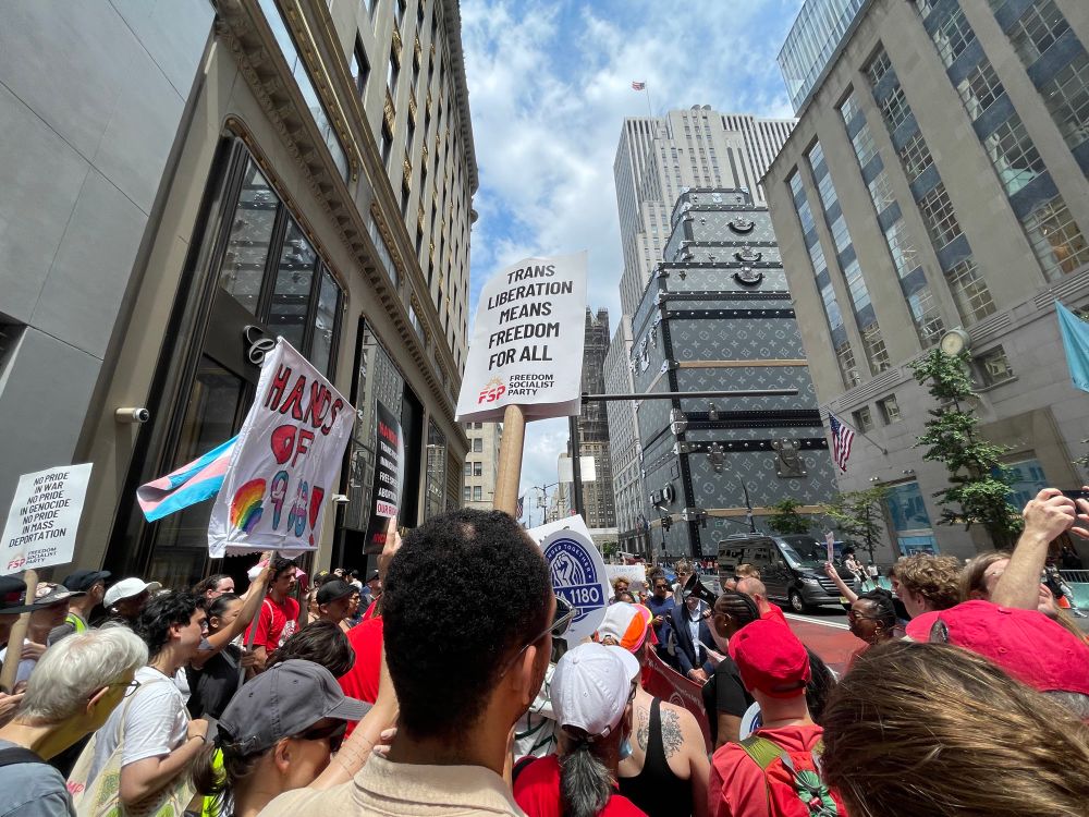 A crowd of protesters stand on 5th Avenue in Manhattan, many wearing red shirts. There is a banner reading “Hands off 988” and a sign that says “Trans liberation means freedom for all.”