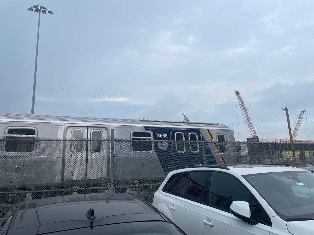 A new subway car (R211A number 3565) sits behind a chain link fence and parked cars in Sunset Park. Cranes, a light pole, and a cloudy sky appear in the background.