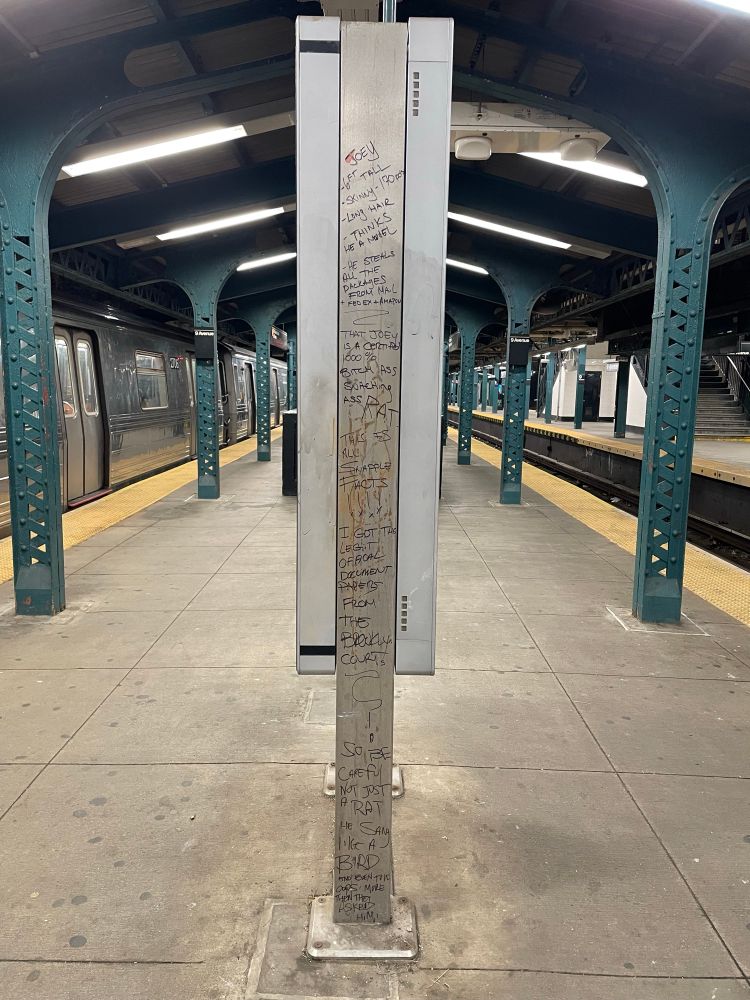A photo of an NYC subway platform. The side of a digital display is covered top-to-bottom in a long sharpie message about a man named Joey who allegedly thinks he’s a model, steals deliveries, and tells the police more than they even want to know.