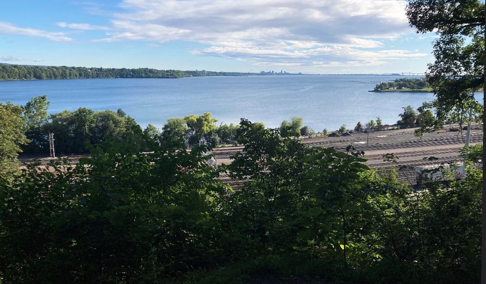 A view of a large lake with railroad tracks in the foreground and trees and a city in the distance. 