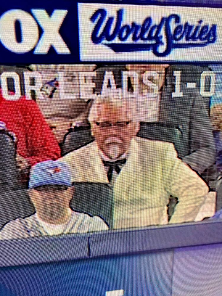Image shows a white man wearing a white suit and wearing glasses sitting behind home plate at the World Series. He looks just like Col Sanders. 