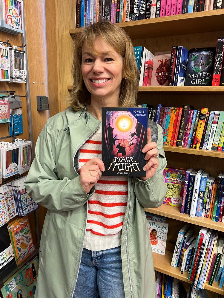 Woman smiling and holding up a spooky middle grade book in front of bookshelves