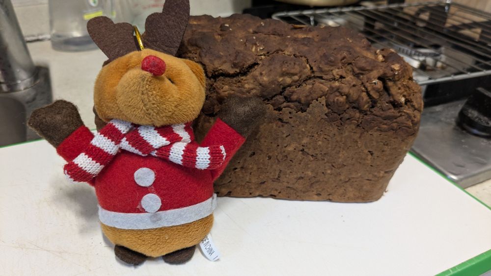 A small stuffed reindeer with a red nose, red coat, and red and white striped scarf standing in front of a loaf of brown bread