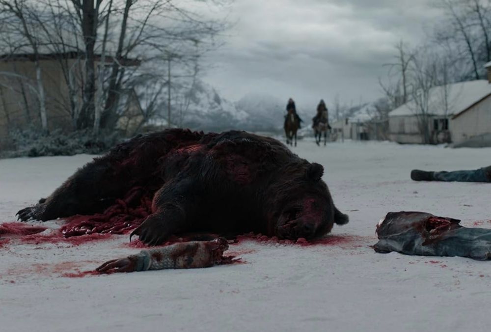 A bloody dead bear in the snow surrounded by zombie body parts with two humans on horseback in the distance