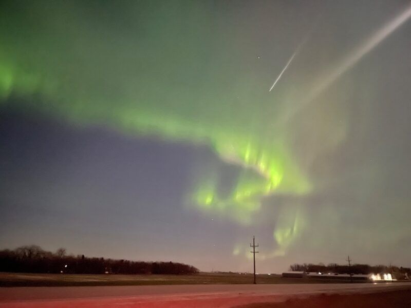 Green aurora swirls above a rural horizon with a bright meteor streaking through the night sky.