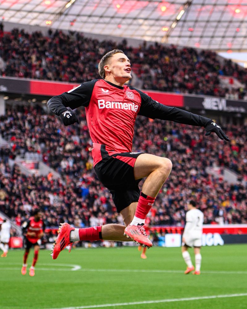 Das Bild zeigt den Fußballspieler Florian Wirtz von Bayer 04 Leverkusen in einem emotionalen Moment während eines Spiels. Er befindet sich in der Luft, offenbar nach einem Tor oder einer entscheidenden Aktion, mit ausdrucksstarker Körpersprache. Wirtz trägt das Heimtrikot seines Vereins: ein rot-schwarzes Jersey mit dem Sponsorenlogo „Barmenia“ auf der Brust, dazu schwarze Shorts, rote Stutzen und rote Fußballschuhe. Schwarze Handschuhe runden sein Outfit ab.

Im Hintergrund ist ein voll besetztes Stadion zu sehen, die Tribünen sind dicht mit Fans gefüllt. Auf dem Spielfeld erkennt man weitere Spieler beider Mannschaften, die Szene wirkt lebendig und spannungsgeladen. Die Körperhaltung von Wirtz – erhoben, dynamisch, mit leicht angewinkelten Armen – vermittelt Freude, Energie und sportliche Leidenschaft.