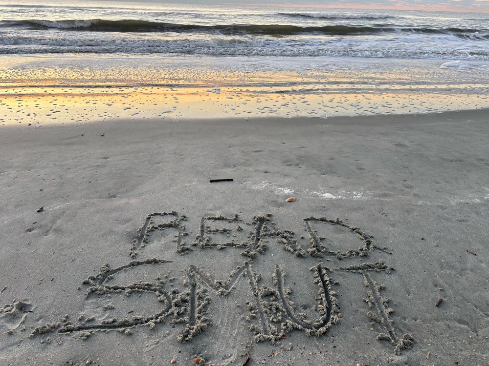 Ocean waves in the background and written in the sand is “Read Smut” 