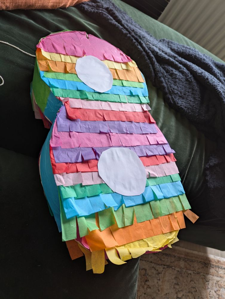 A homemade piñata in the shape of a number eight. It has colourful strips of tissue paper stuck across it. White paper circles form the two holes in the number