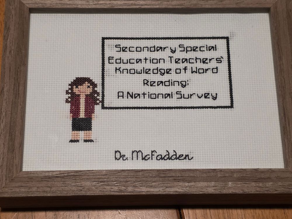 Cross stitched stitched canvas showing a cartoon character representing Kim defending her dissertation titled Secondary Special Education Teachers' Knowledge of Word Reading: A National Survey 