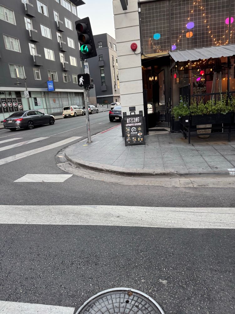 A sidewalk corner curb on fourth and Main with no ADA ramp. Opposite corner.