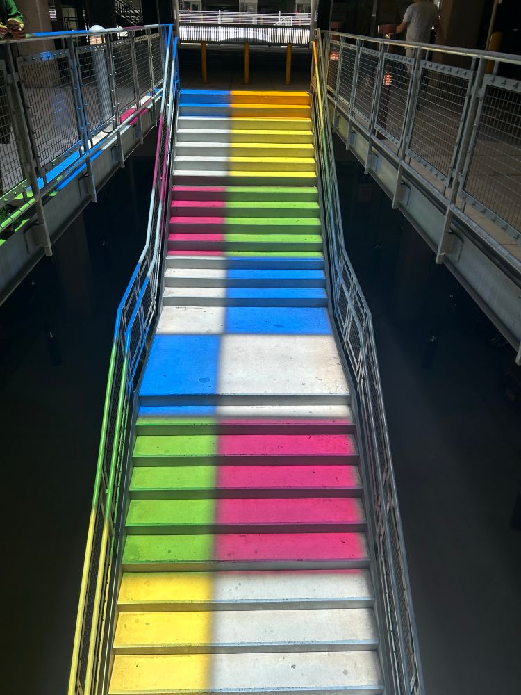 An image of the solar colored panels casting blocked lighting on a concrete staircase with iron handrails. Passersby walk past the staircase toward the entrance of San Antonio International Airport.