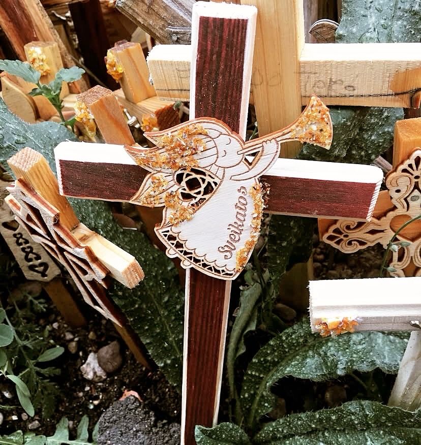 A small wooden folk cross, with an amber encrusted angel in the center, playing a trumpet. The word Sveikatos, which means I wish you health, is written on the angel’s gown.