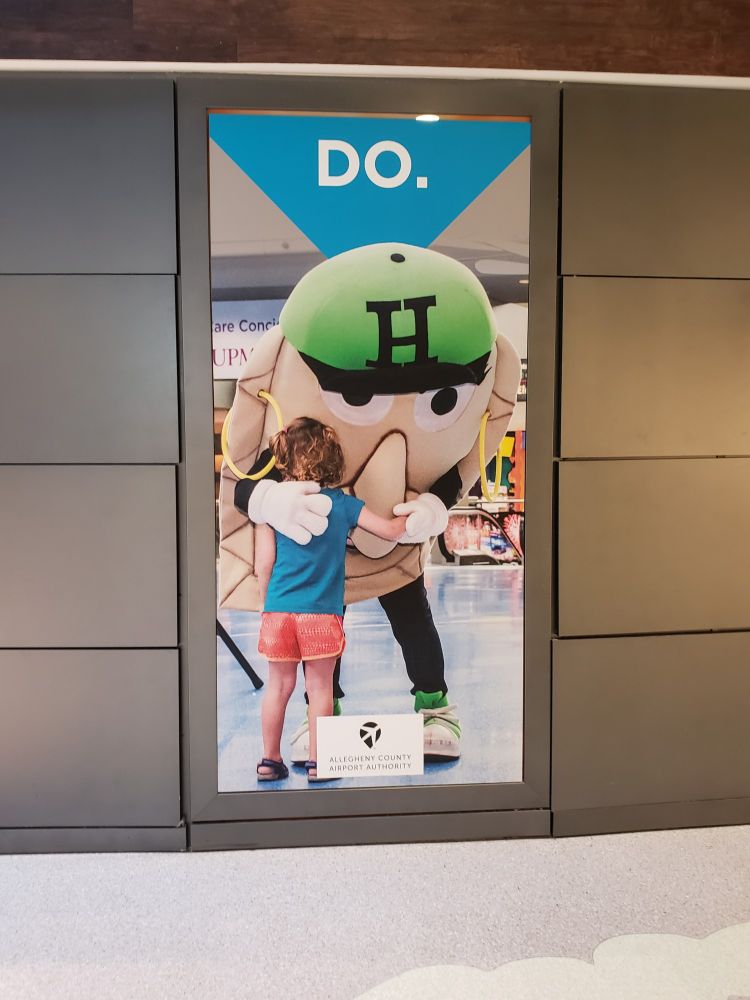 Advertisement of a child hugging a person in a pierogie costume. It is displayed at the Pittsburgh Airport. A pierogie is an eastern European dumpling with potato or other fillings on the inside.