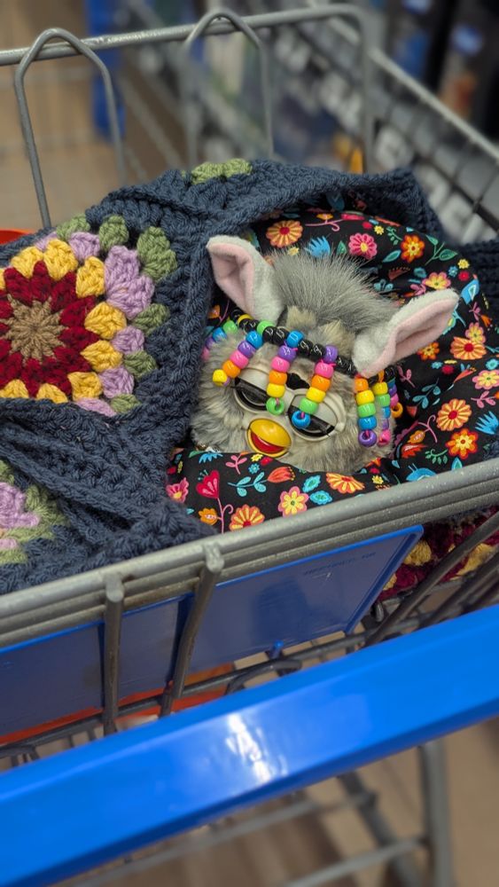 A grey wolf 1998 furby sits ina handmade crochet bag nestled in the baby seat of a shopping cart