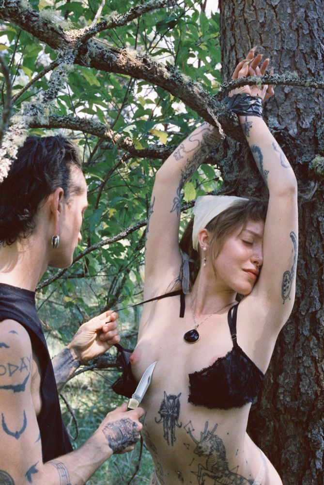 Beau expresses despair as she presses her face against her arm. Her wrists are duct tape bound over her head and are stuck in between a tree branch. Molly traces a knife down her chest as she pull on Beau’s bra, which has been sliced in half by Mollys knife. Their scene takes place in between a multi branched tree.