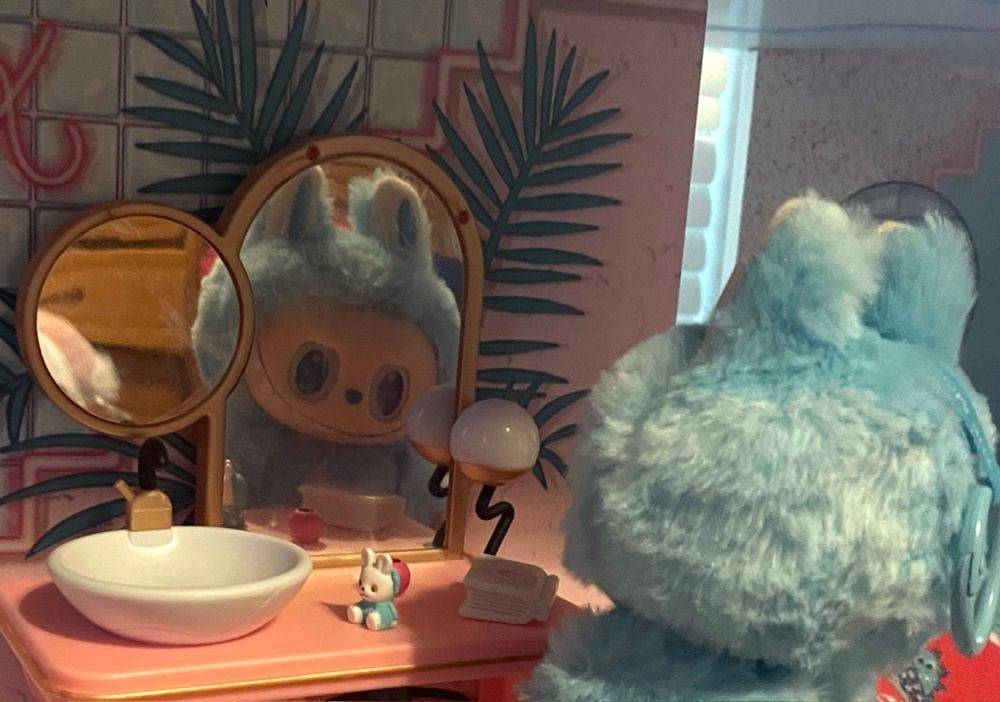 A blue Labubu stares at their reflection in a dollhouse mirror.