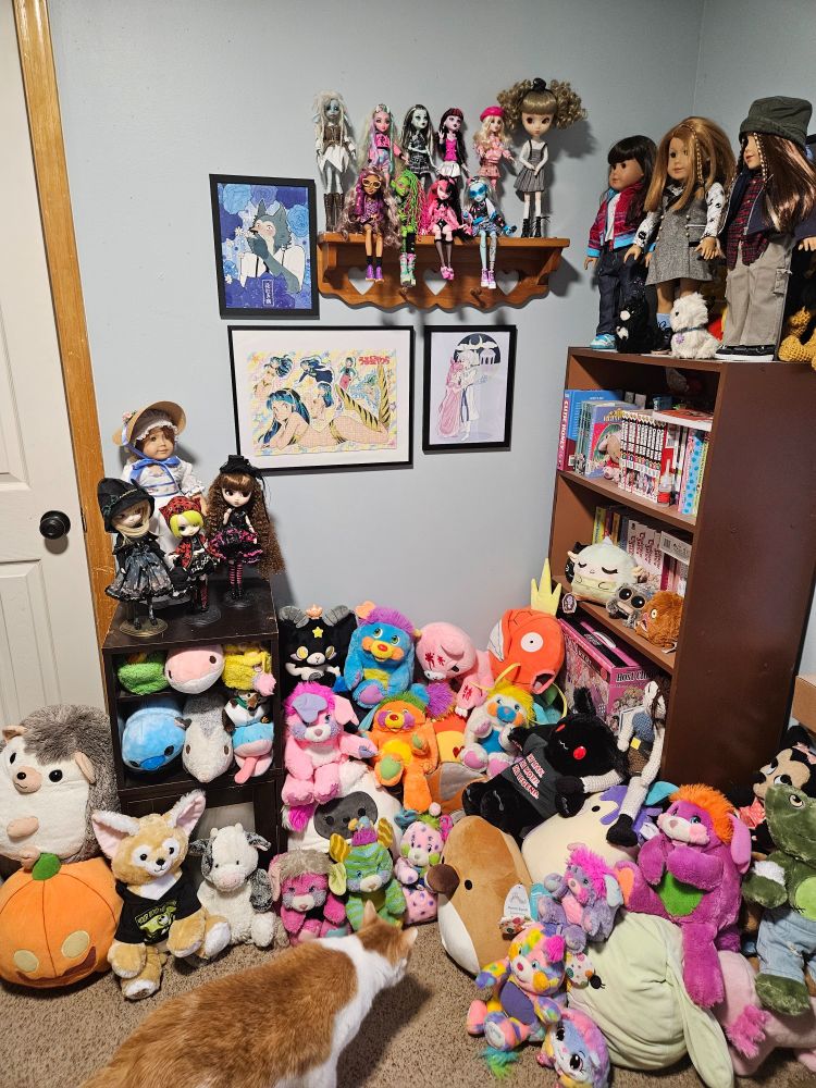 a cartoonish corner of a room with plushies piled on the floor, a book case full of manga with three AG dolls up on top, a shelf on the wall with MH and a Pullip doll, framed anime prints on the wall beside and below that, and a nightstand with a Felicity AG doll and two Pullips and a Dal. My orange and white cat, Meelo, sniffs the pile of plush.