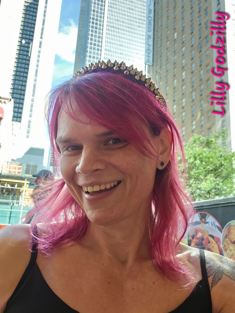 Lilly Godzilly close up with buildings behind her

She has a blsck dress, pink hair and a metal studded headband 