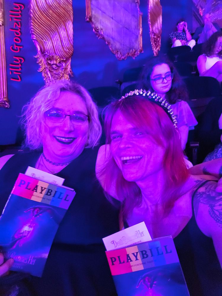 Lilly and Scotia are both wearing black dresses, and lillys dress has skulls in lepoard print on the skirts

 Scotia has blonde hair and Lilly has pink hair adorned with an amazing black headband with crome spikes all over it

The picture has a purple hue from the lights and they are holding up the playbills for the show 

Image is signed Lilly Godzilly