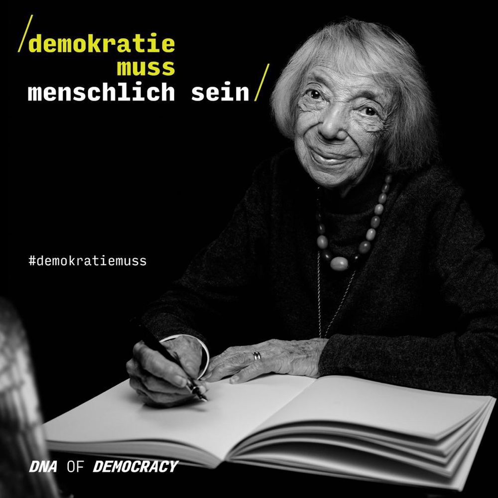 Foto von Margot Friedländer in schwarz-weiß, wie sie bei der Kampagne DNA of Democracy in das Buch schreibt, was das Grundgesetz symbolisieren soll. 