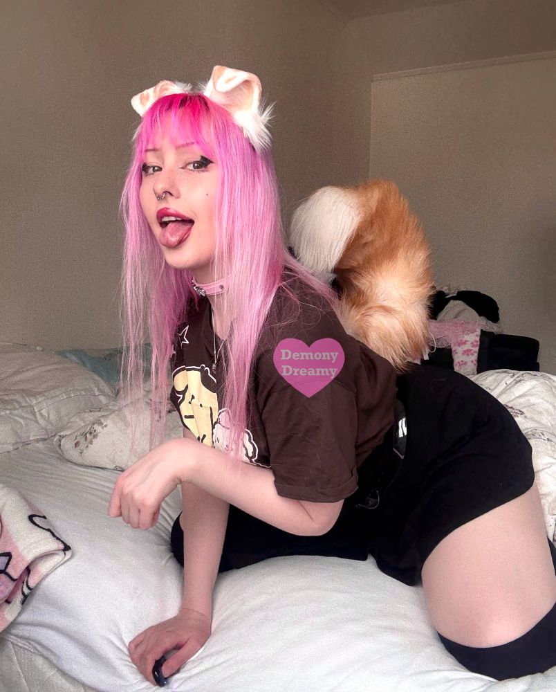 I’m sitting on all fours on a bed, with one hand held up making a pawing motion. I’m sticking my tongue out in a panting motion and looking at you with a smile. I’m wearing white and light brown spotted puppy ears and a tail, a pastel pink collar, a silver necklace with a heart pendant, a dark brown T-shirt with Pompompurin and Macaroon printed on it, a black skirt and black thigh high socks.