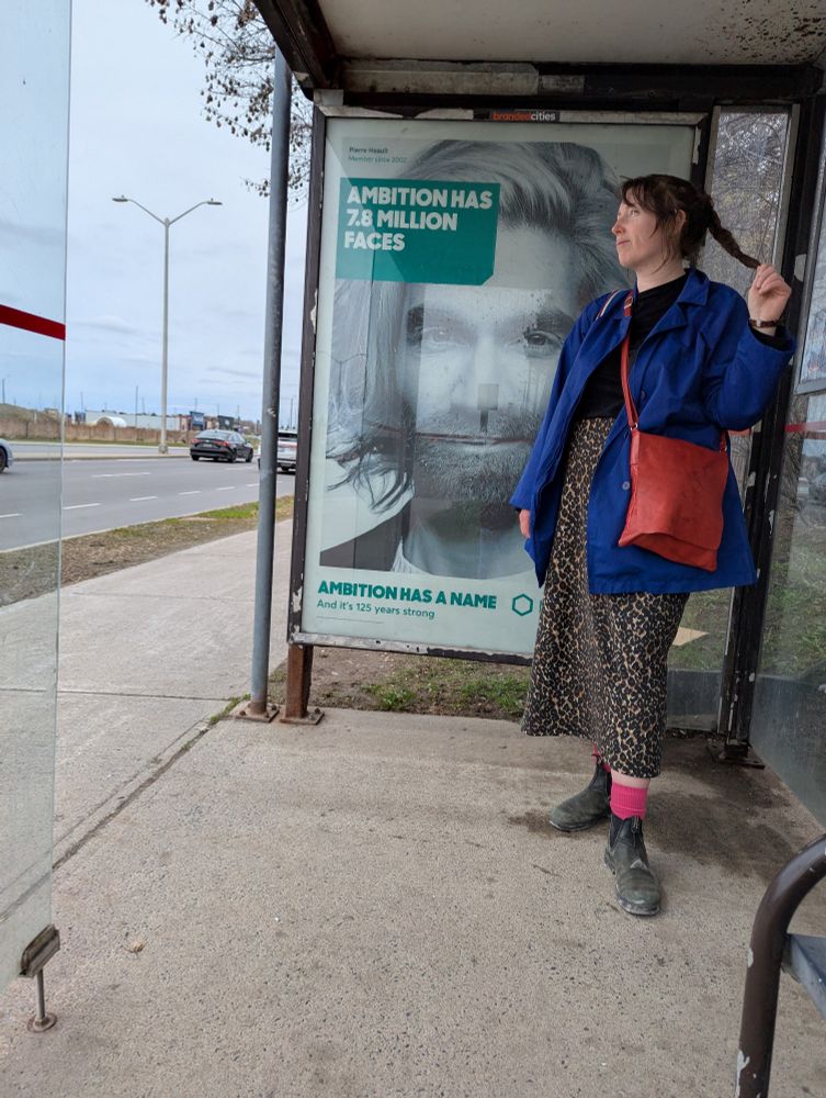 A tall white woman stands in a bus shelter, looking off to the left. Her long dark hair is in a braid. She is wearing a blue coat, a red crossbody bag, pink socks, and Blundstone boots. The highlight of the photo is the leopard print skirt which she made.