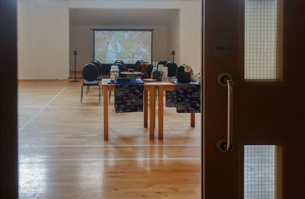 a distant view from doorway of the event setup, projection screen with speakers, lines of chairs, and tables for food and drinks. 
