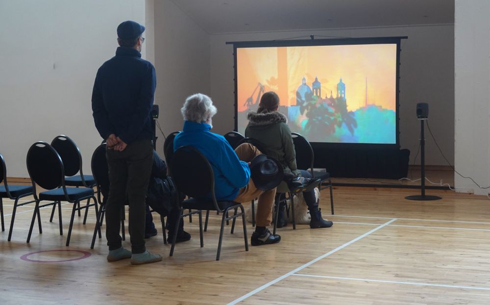 a group of people sitting on chairs and standing, with a white dog lying next to them, watching projected films in a community hall. A pair of speakers visible on the sides of the screen.
